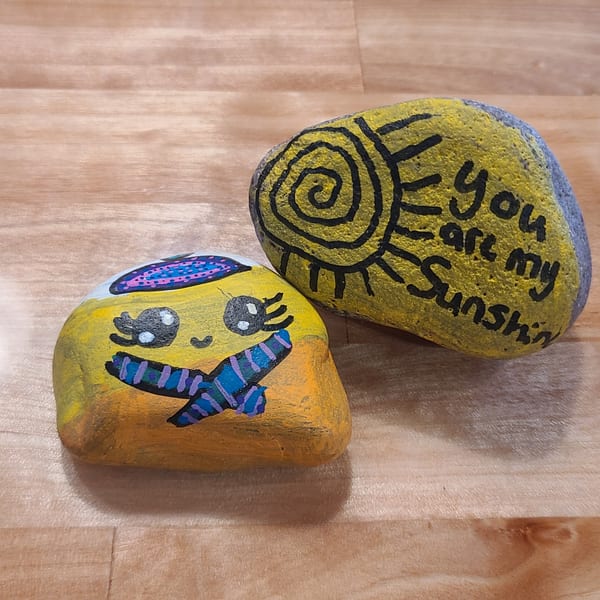 Two painted rocks on a wooden surface, one with a face and scarf, the other with "You are my Sunshine".
