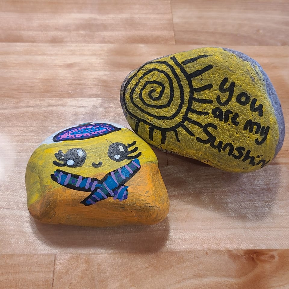 Two painted rocks on a wooden surface, one with a face and scarf, the other with "You are my Sunshine".