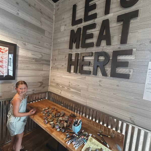 Girl painting rocks at the Happy Little Rock station under the Lets Meat Here sign