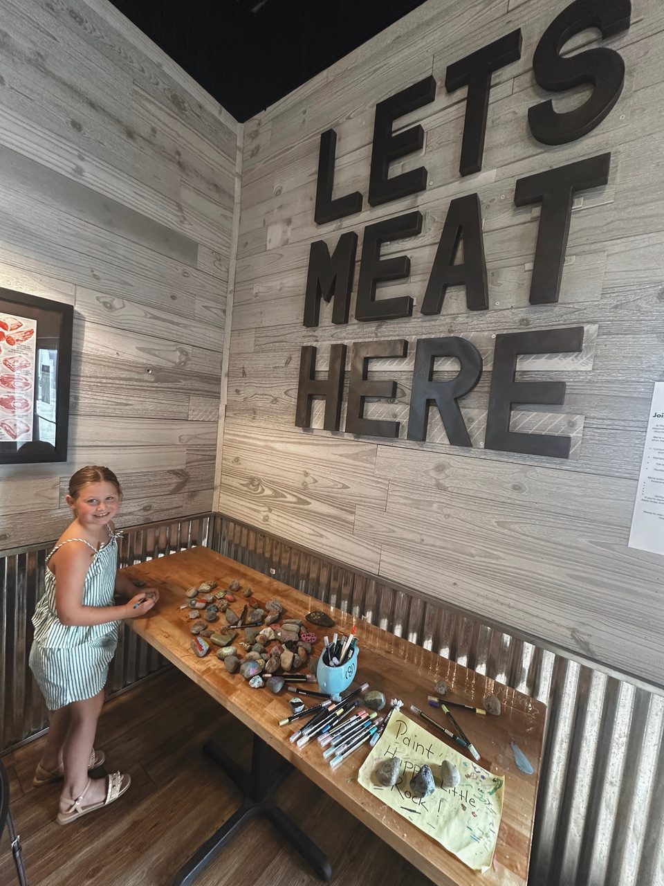 Girl painting rocks at the Happy Little Rock station under the Lets Meat Here sign